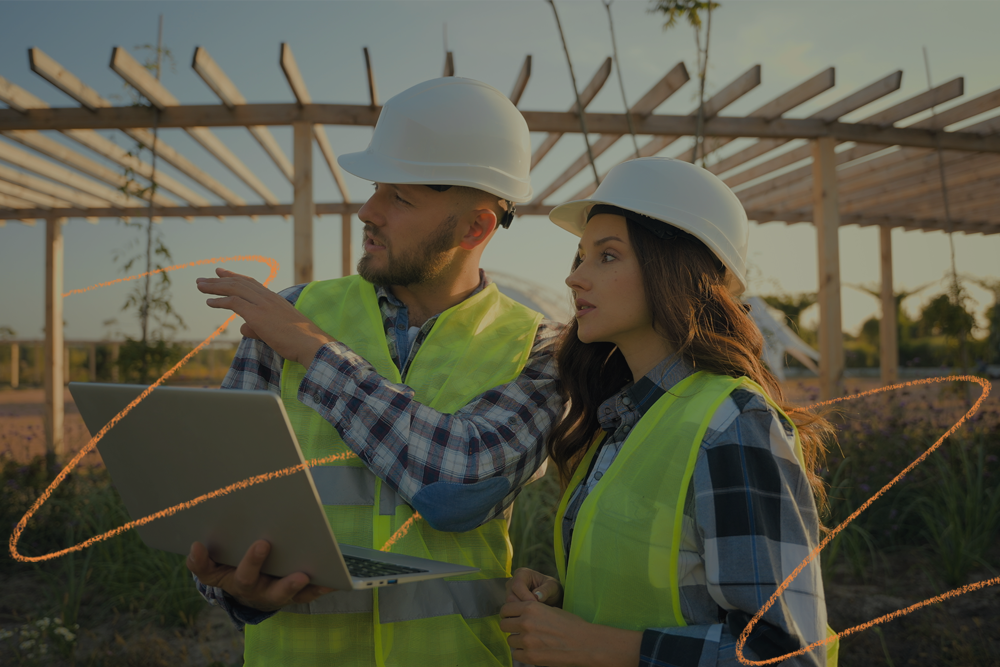 Dois profissionais da construção civil, usando capacetes e coletes refletivos, analisam um projeto em um notebook em meio a uma obra ao ar livre. A imagem ilustra a aplicação prática de anúncios digitais na construção civil, destacando como a tecnologia e o marketing estão cada vez mais presentes nas decisões estratégicas do setor.